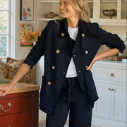 A woman stands in a bright home office wearing a tailored navy double-breasted blazer with matching trousers, a white top, and tan loafers. She is leaning against a wooden desk next to a window, with shelves and a patterned rug in the background.