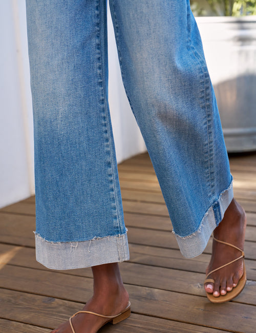 Close-up of a person wearing light blue wide-leg cuffed jeans with a raw frayed hem, paired with tan flat slide sandals on a wooden deck., view 3