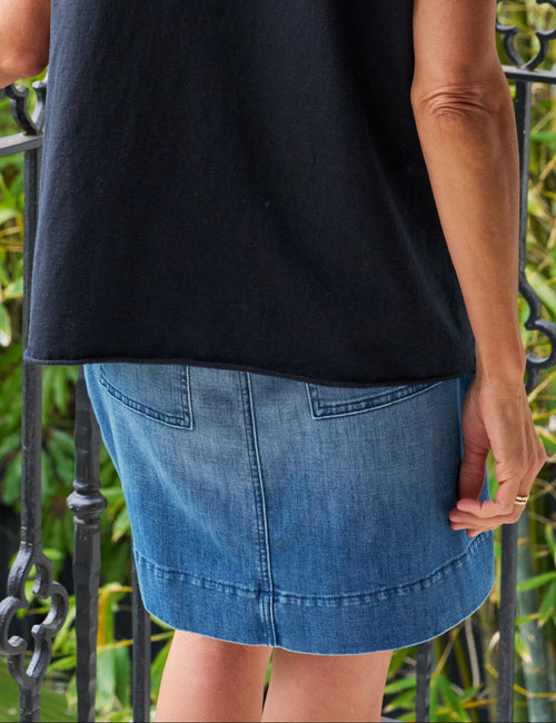 Woman wearing a navy sleeveless top and denim skirt standing on a balcony with greenery in the background, view 4