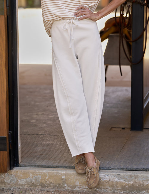 A person wearing loose, barrel-leg cream or off-white drawstring pants with a striped top and tan lace-up shoes, standing in a doorway., front view