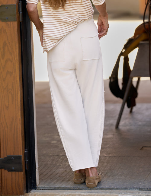 A person shown from the back wearing loose white barrel-leg pants with back patch pockets, a cream and tan striped top, and tan slip-on shoes, standing in a doorway., alternate view