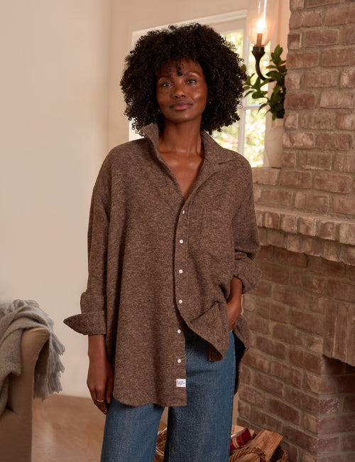 A woman wearing a brown textured button-up shirt and blue jeans, standing in a cozy room with a brick fireplace and a window in the background., front view