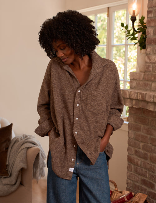 A woman wearing an oversized brown button-up shirt and blue jeans, standing in a cozy room with a brick fireplace and a window in the background., alternate view