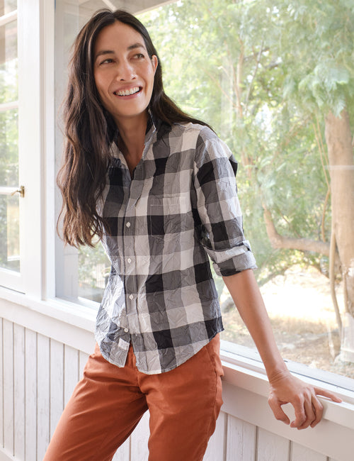 A woman smiling while wearing a black and white checkered button-down shirt and rust-orange pants, leaning on a white porch railing with trees visible in the background., front view