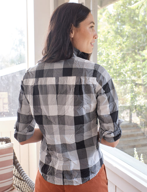A woman wearing a black, white, and gray buffalo plaid button-down shirt, shown from the back as she looks out a window., alternate view