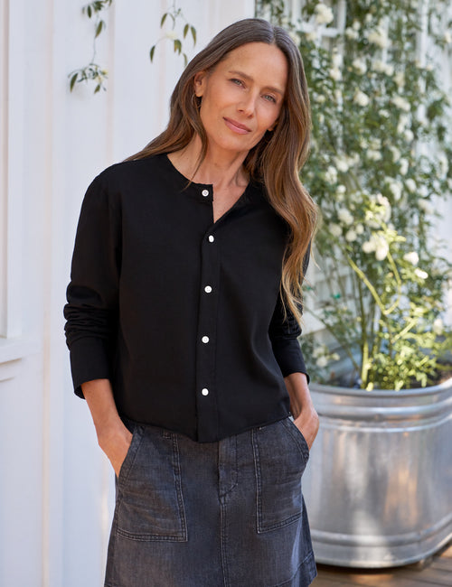 Woman wearing a black button-up shirt and denim skirt standing outdoors with plants in the background, front view
