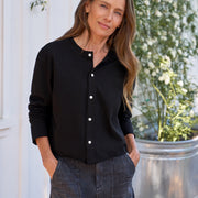 Woman wearing a black button-up shirt and denim skirt standing outdoors with plants in the background
