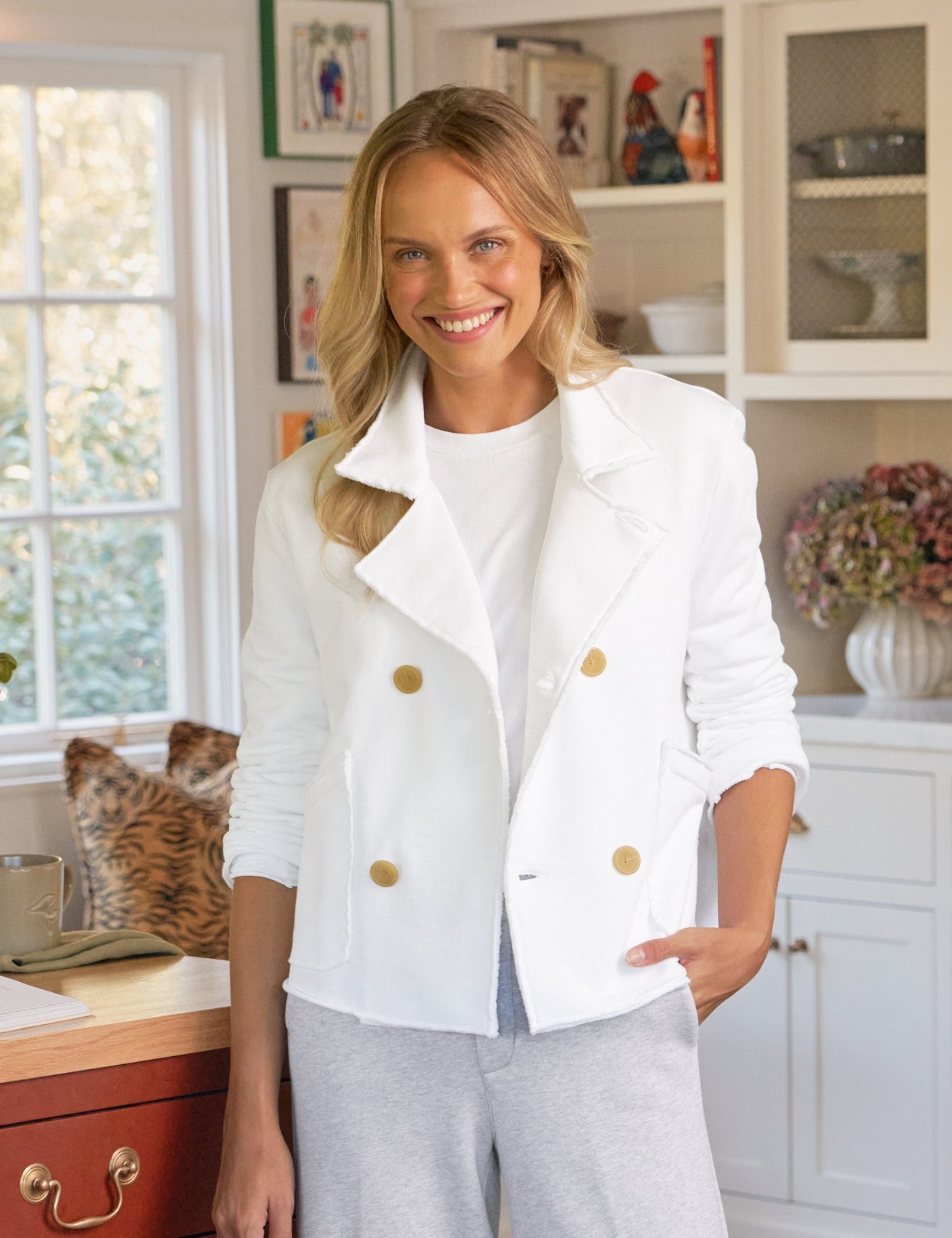 Woman in a white jacket and light gray pants standing in a kitchen., front view