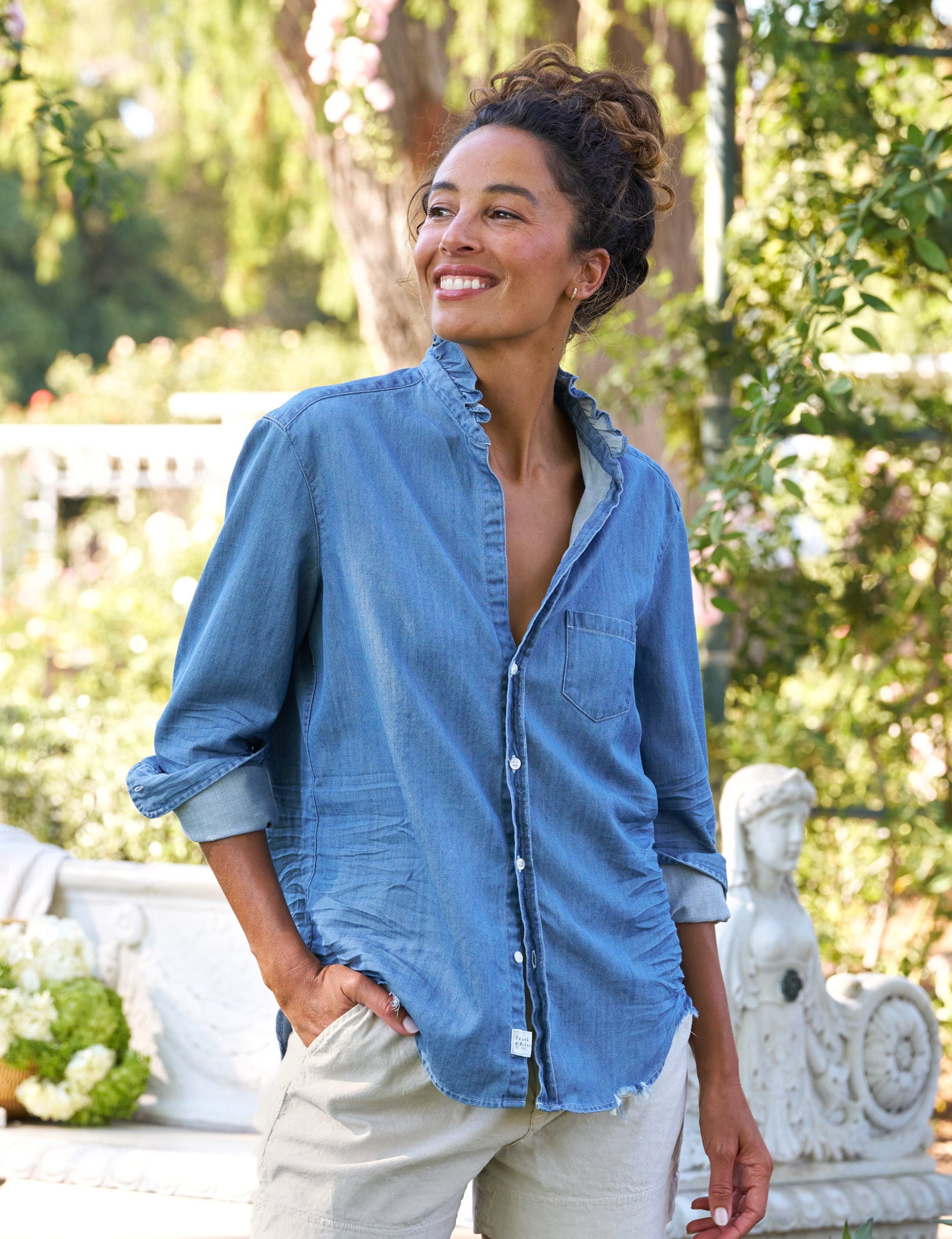 Woman wearing a blue shirt and beige shorts standing outdoors with greenery in the background, front view