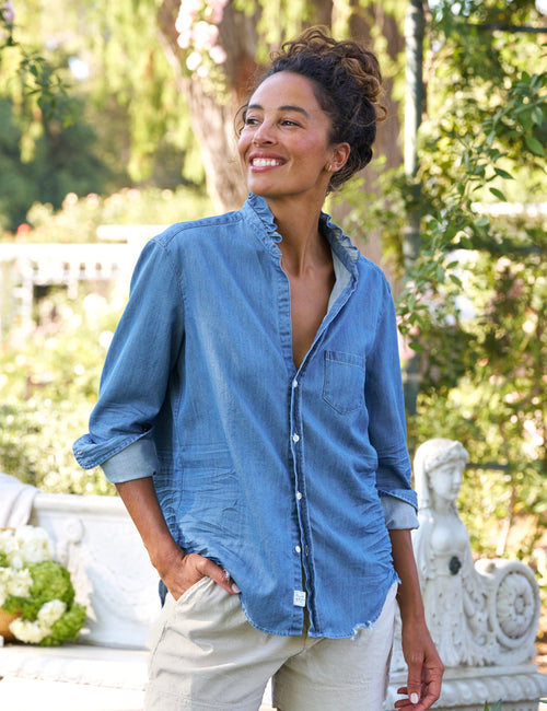 Woman wearing a blue shirt and beige shorts standing outdoors with greenery in the background, front view