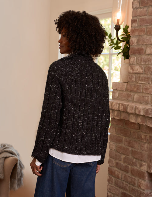 A woman with curly dark hair is shown from the back wearing a black textured knit cardigan with silver sequins, layered over a white shirt and blue jeans, standing in a room with a brick fireplace., alternate view