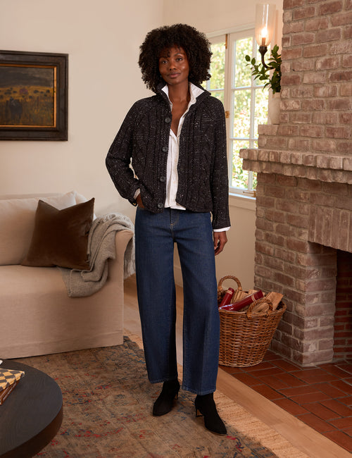 A woman stands in a cozy living room wearing a dark textured knit cardigan over a white collared shirt, paired with blue wide-leg jeans and black ankle boots., view 4