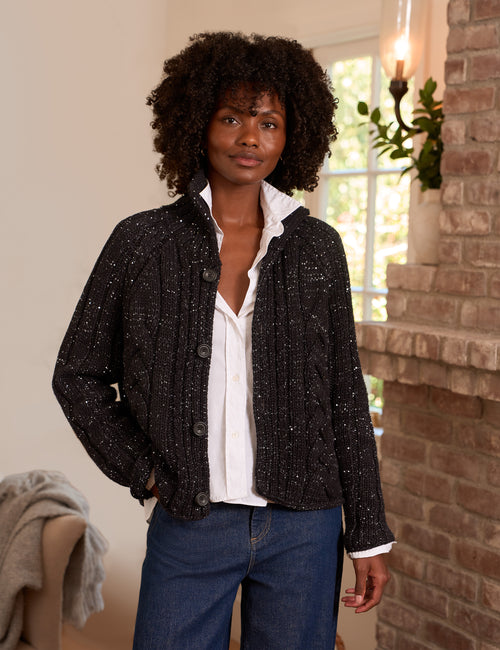 A woman with curly black hair stands indoors wearing a black sequin knit cardigan with a white button-up shirt and blue jeans. The setting features a warm brick fireplace on the right and a neutral wall on the left., front view