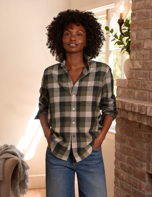 A woman with curly dark hair stands indoors wearing an oversized green and beige plaid button-up shirt with blue jeans, posed in front of a brick wall and window., front view