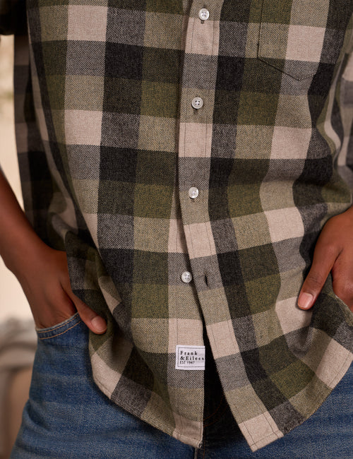 Close-up of a person wearing a plaid button-up shirt in olive green, beige, and black, with hands in blue jeans pockets. A small 'Frank & Eileen' brand label is visible near the hem., view 3