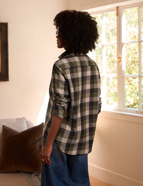 A woman with curly dark hair stands in profile in a sunlit room, wearing an oversized green and beige plaid button-up shirt with blue jeans., alternate view
