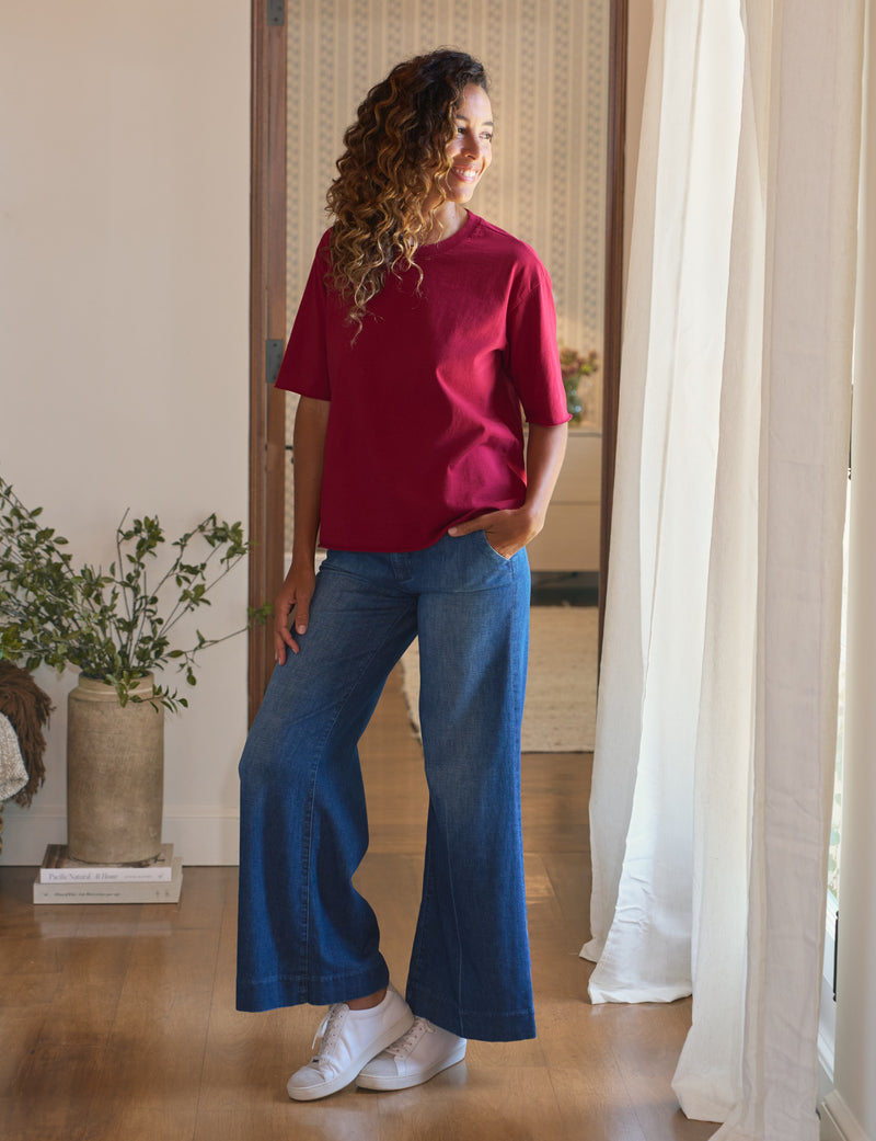 Woman wearing a red shirt and blue jeans standing in a room with a plant and white curtains.