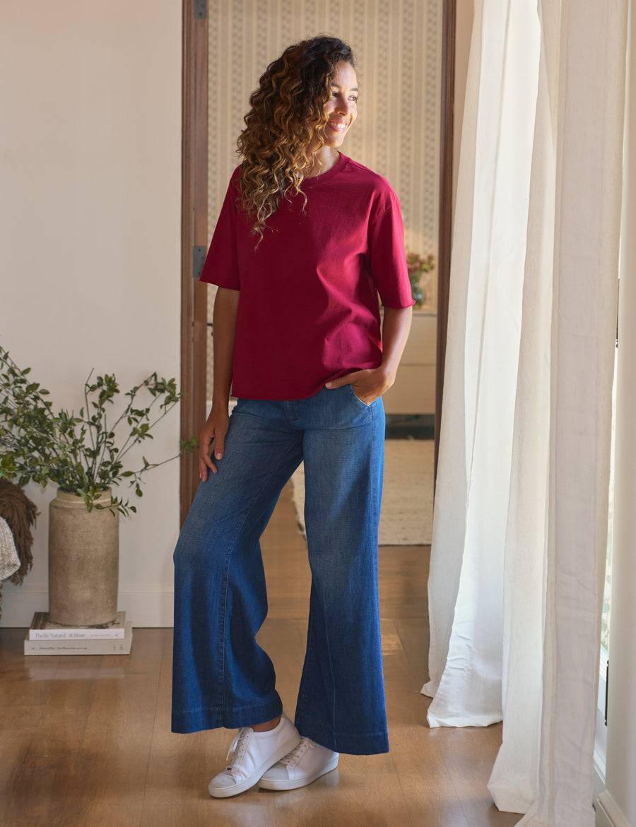 Woman wearing a red shirt and blue jeans standing in a room with a plant and white curtains.