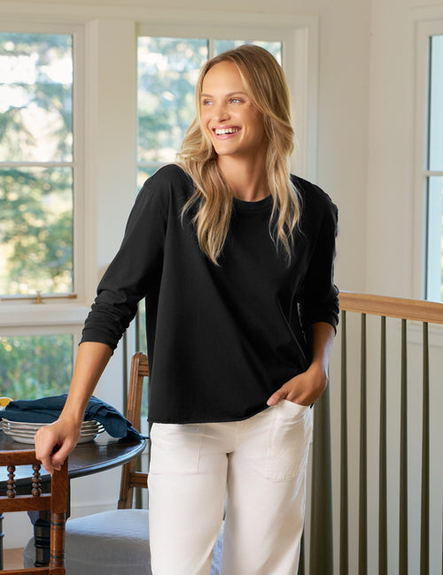 Woman standing in a room with large windows, smiling and wearing a black top and white pants., front view