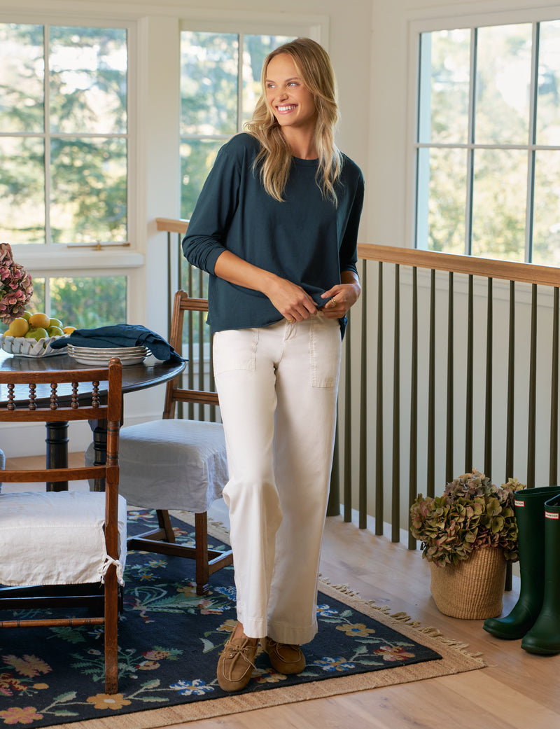 Woman standing in a room with large windows, wearing a dark blue tee and white pants.