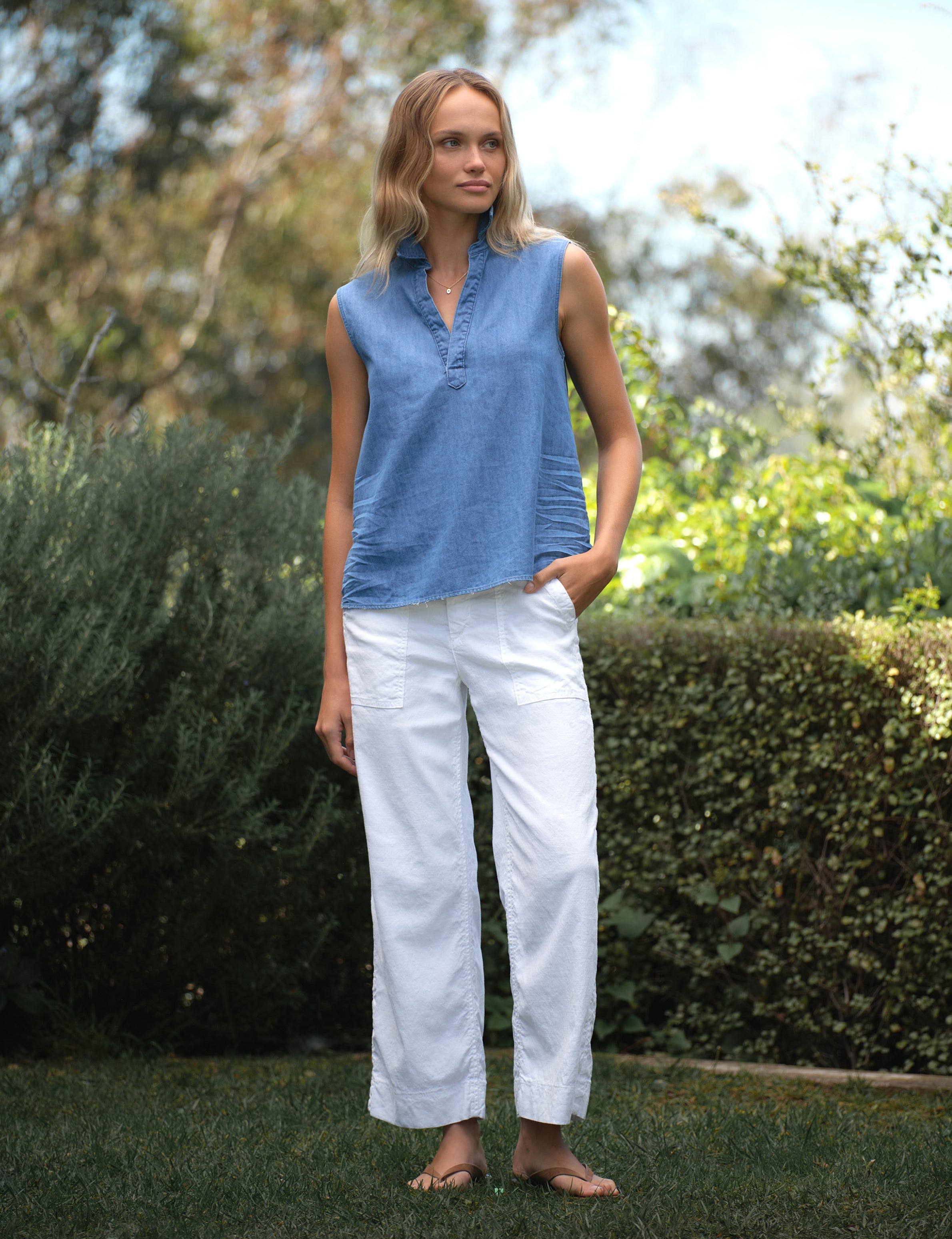 Woman wearing a blue sleeveless top and white pants standing outdoors with greenery in the background, view 5