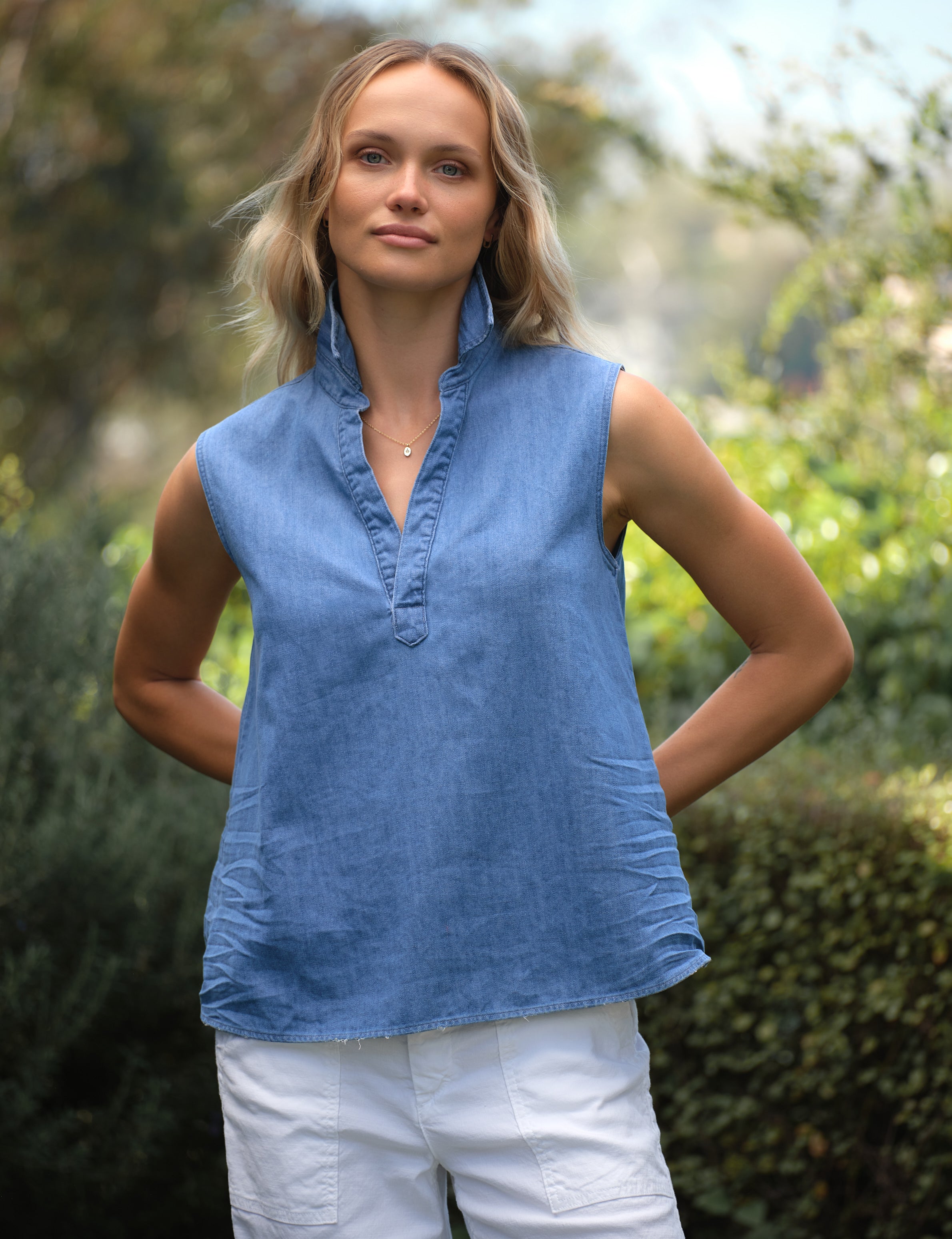 Woman wearing a blue sleeveless top and white pants standing outdoors with greenery in the background, front view