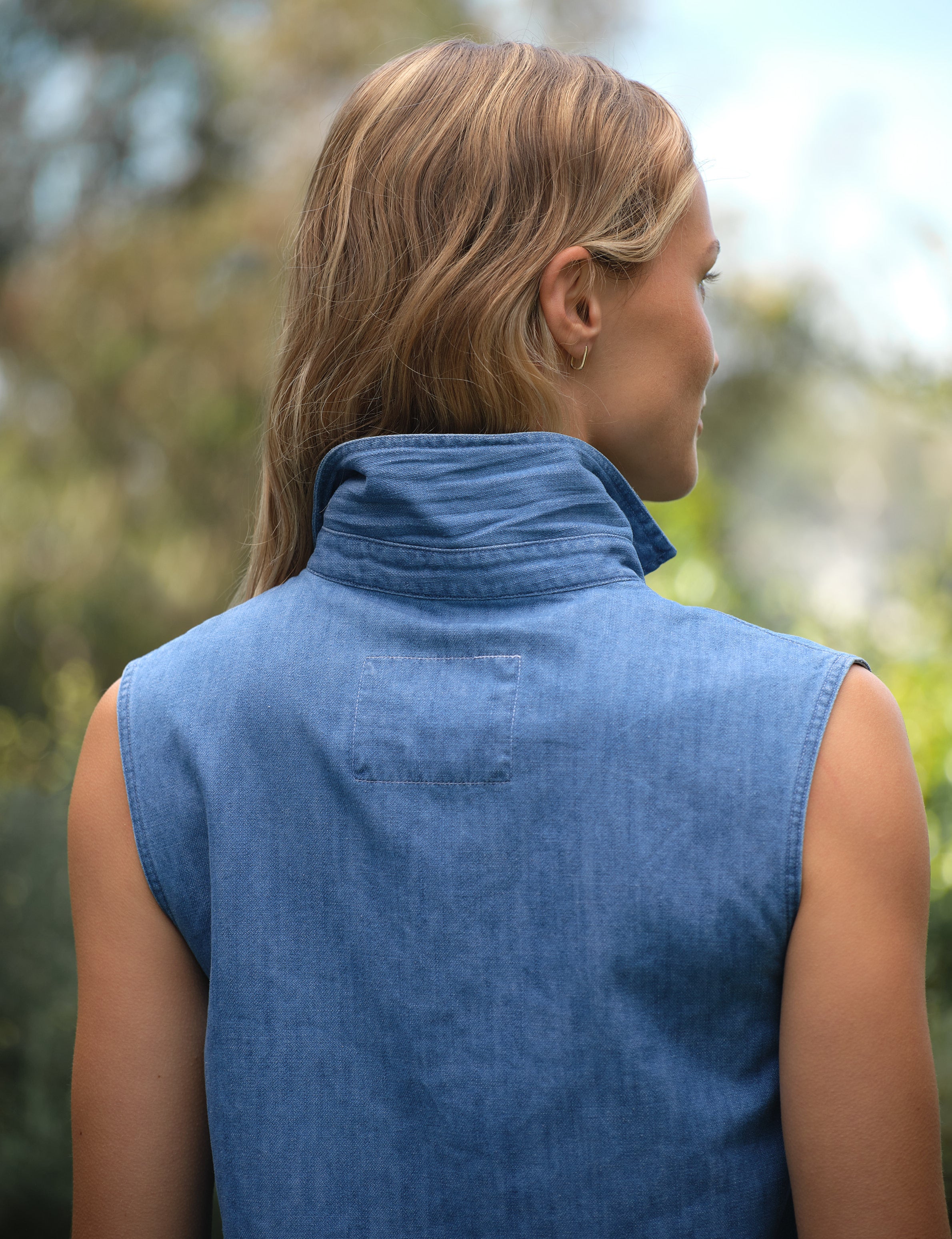 Person wearing a blue sleeveless denim shirt outdoors with blurred greenery in the background, alternate view