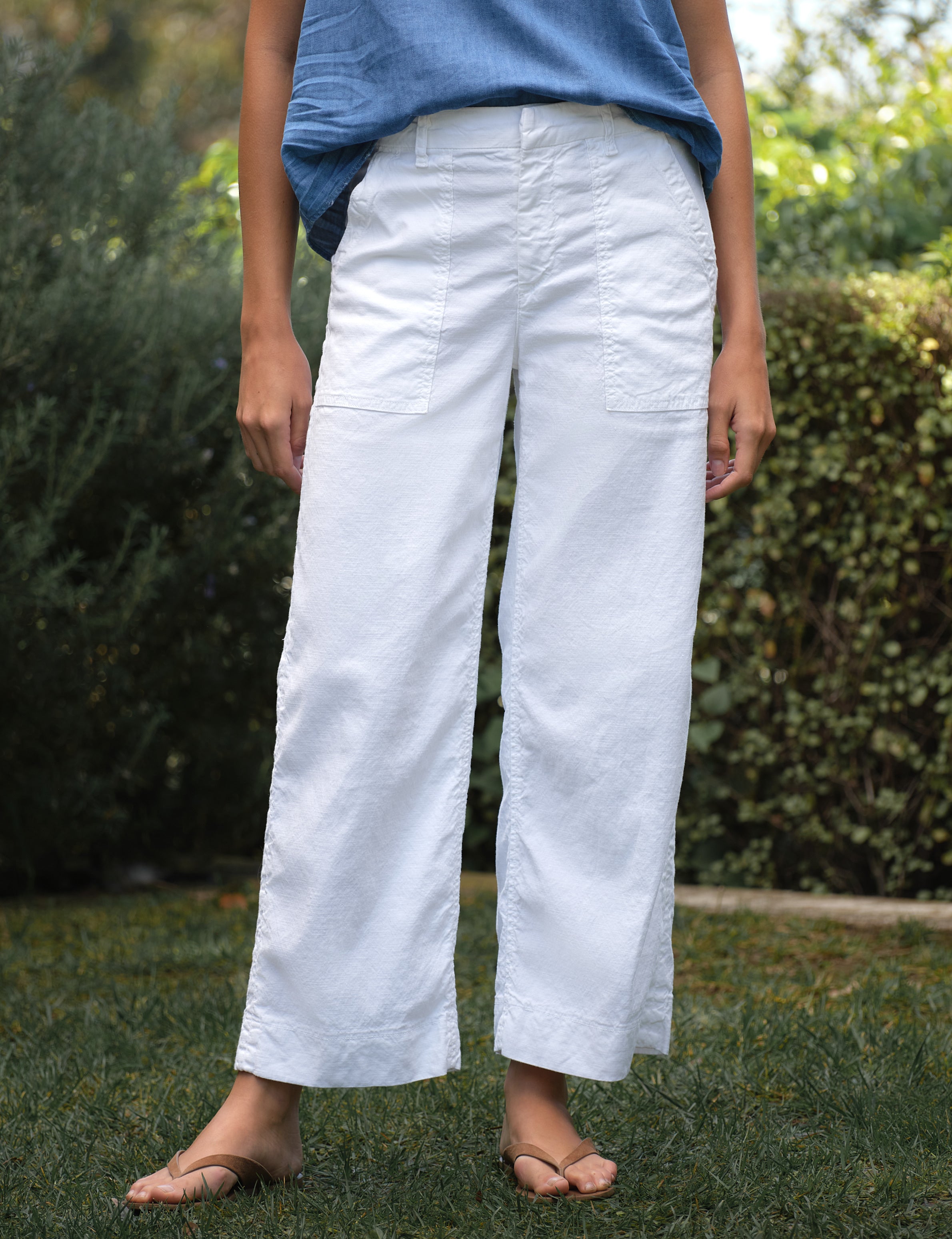 Person wearing white pants and a blue top standing outdoors with greenery in the background, front view