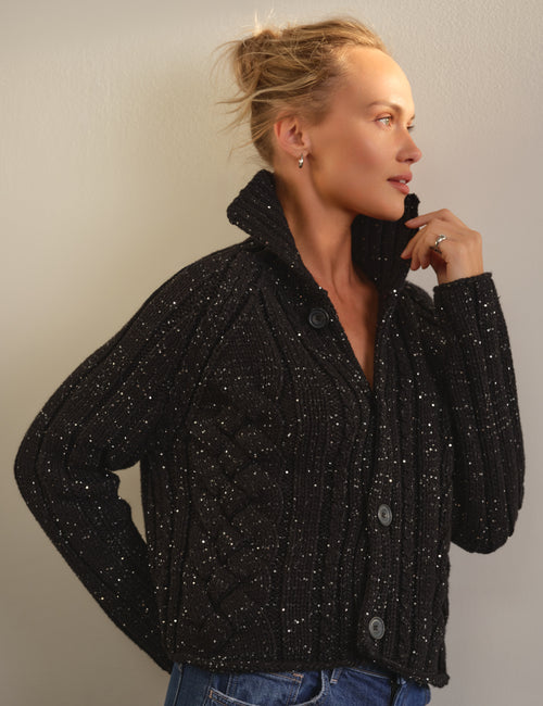 A woman wearing a black cable-knit cropped cardigan sweater with silver sequins, styled with blue jeans against a plain light gray wall., view 5