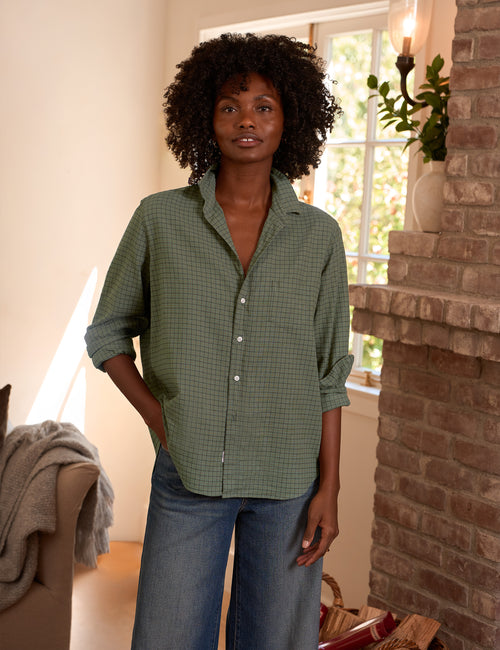 A woman with curly black hair stands indoors wearing an olive green and brown micro-check button-down shirt with blue jeans. The setting features a brick fireplace, a window, and a neutral-toned armchair., front view