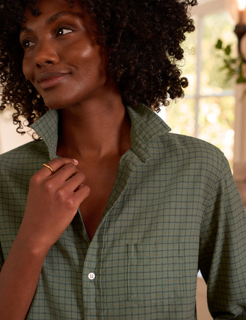 A close-up lifestyle shot of a woman with curly black hair wearing a sage green button-down shirt with a fine brown grid pattern. She is looking off-camera with a soft smile, and the background is a softly blurred indoor setting., view 3
