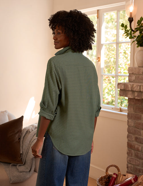 A woman with curly black hair is shown from the back wearing a sage green and brown checkered button-down shirt with rolled-up sleeves, paired with blue jeans, in a warm, sunlit room with a brick fireplace., alternate view