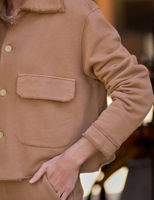 Close-up of a person wearing a tan utility jacket with a collar, front flap pocket, and light buttons, with one hand in a pocket., view 3