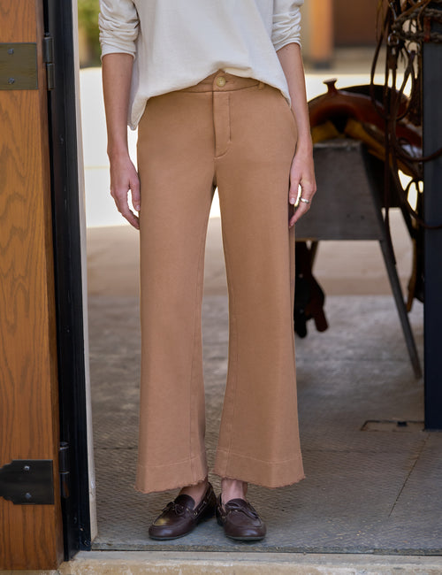 A person wearing tan wide-leg cropped pants with a cream long-sleeve top and dark brown loafers, standing in a doorway., front view
