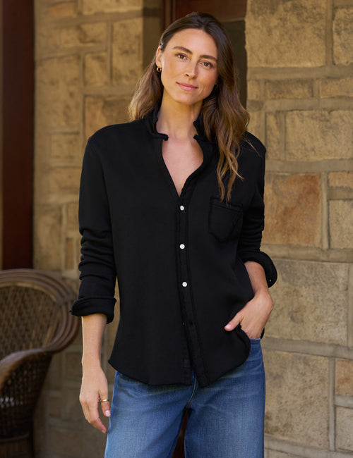A woman wearing a black long-sleeve button-up shirt and blue jeans, standing in front of a stone wall., front view