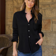 A woman wearing a black long-sleeve button-up shirt and blue jeans, standing in front of a stone wall.