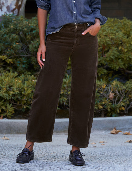 A person wearing dark brown barrel-leg corduroy pants, styled with a blue denim shirt and black shoes, standing outdoors in front of green bushes., front view