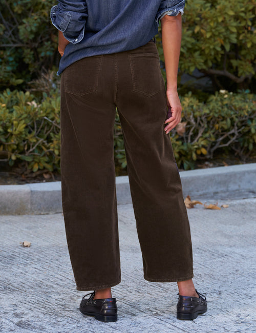 A person shown from the back wearing dark brown barrel-leg corduroy pants, a blue denim shirt, and black shoes, standing outdoors on a paved surface with green bushes in the background., alternate view