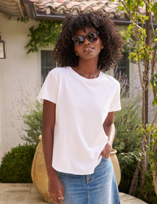 Woman wearing sunglasses and a white t-shirt in front of a house with greenery., front view