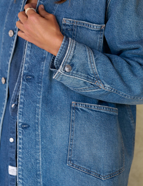 Close-up of a person wearing a medium-wash blue denim button-up shirt jacket with front pockets and a visible hand adjusting the collar., view 4
