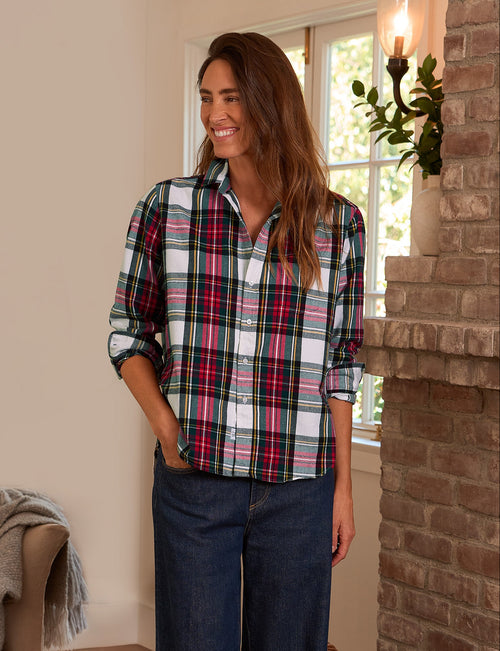 A woman wearing a long-sleeve button-up plaid shirt with red, green, black, and white stripes, paired with dark blue jeans, standing in a cozy room with a brick fireplace and window., front view