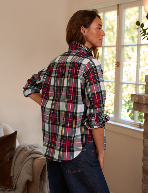 A woman wearing a red, green, and white plaid flannel shirt with rolled-up sleeves, paired with dark blue jeans, standing in a brightly lit room near a window., alternate view