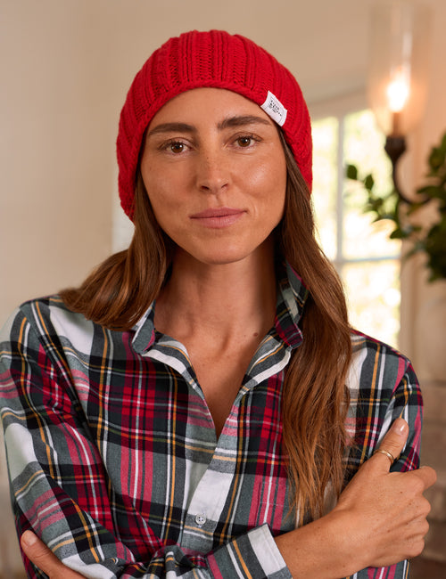 A woman wearing a bright red ribbed knit beanie with a small white brand tag and a red, green, and white plaid button-up shirt, posed indoors with her arms crossed., alternate view