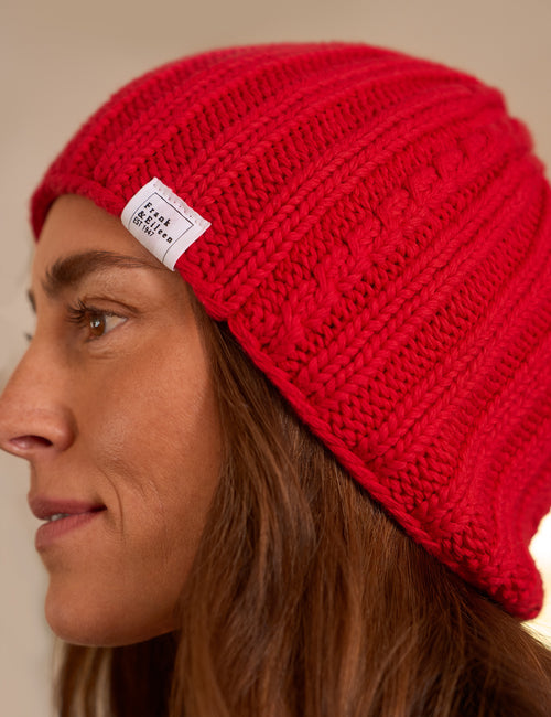 A close-up side profile of a woman wearing a bright red cable-knit beanie with a small white brand label that reads 'Frank & Eileen'., view 3