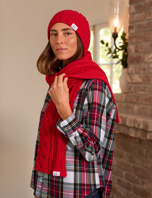 A woman wearing a bright red cable-knit beanie and matching red knit scarf, layered over a red, black, and white plaid flannel shirt. She is posed in front of a brick wall and a window., front view