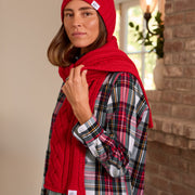 A woman wearing a bright red cable-knit beanie and matching red knit scarf, layered over a red, black, and white plaid flannel shirt. She is posed in front of a brick wall and a window.