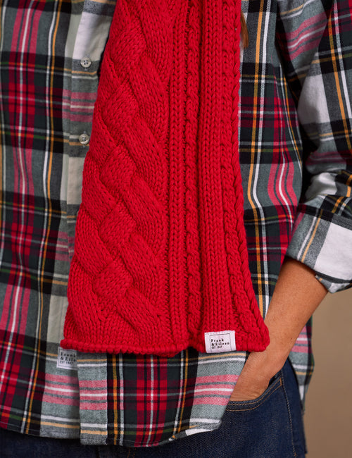A close-up of a person wearing a bright red cable-knit scarf over a red, black, and gray plaid flannel shirt, with a small white brand tag visible on the scarf., alternate view