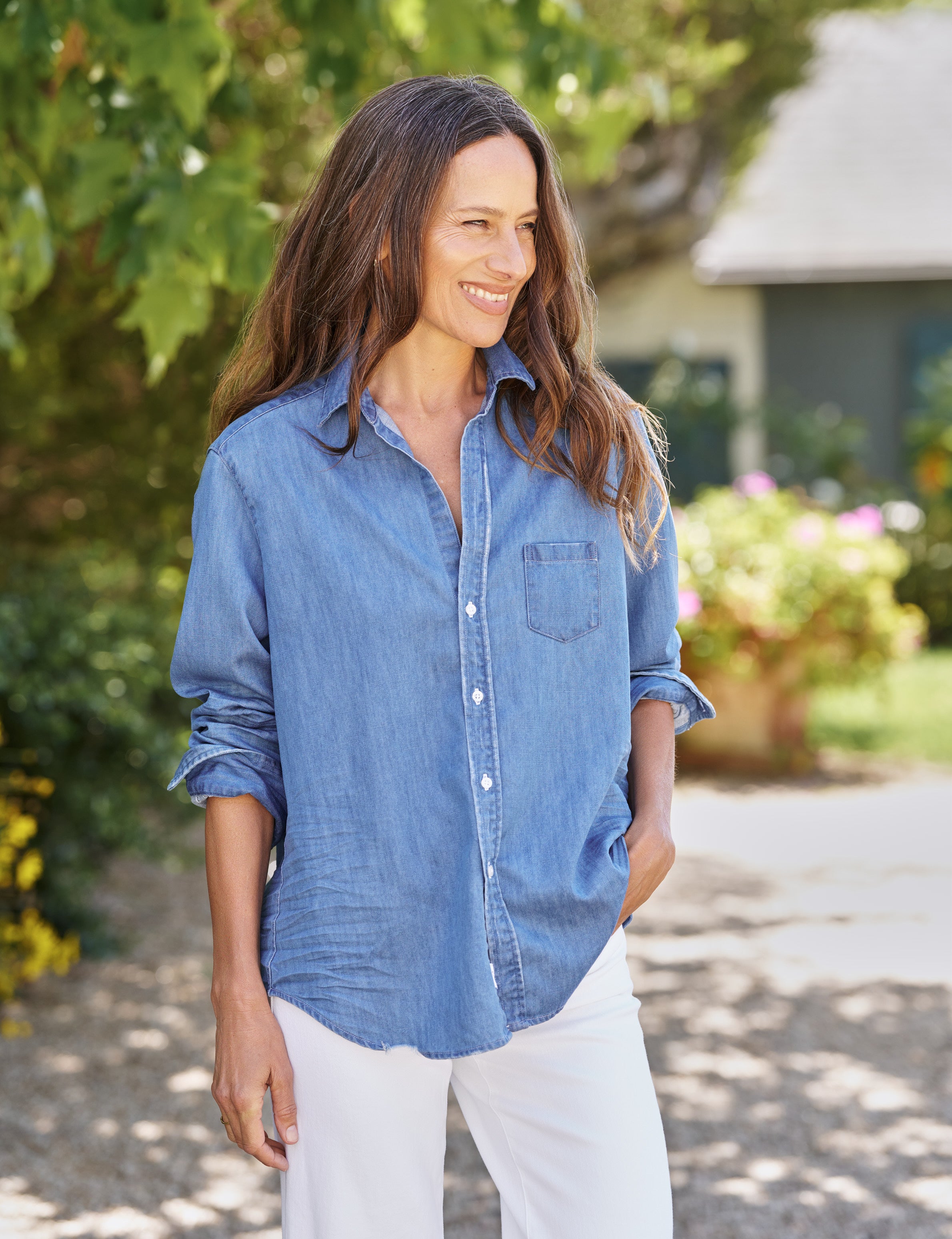 Woman wearing a blue denim shirt and white pants standing outdoors with greenery in the background, alternate view