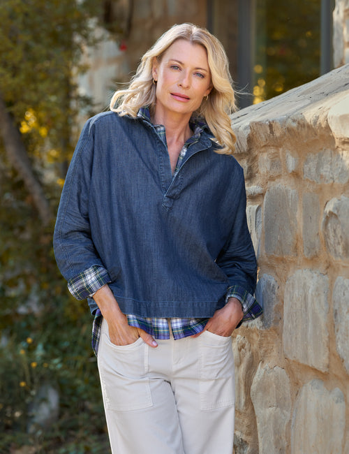 Woman wearing a blue pullover with a plaid shirt underneath, standing against a stone wall., view 5