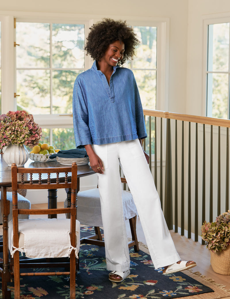 Woman in a blue shirt and white pants standing in a room with a dining table and chairs.